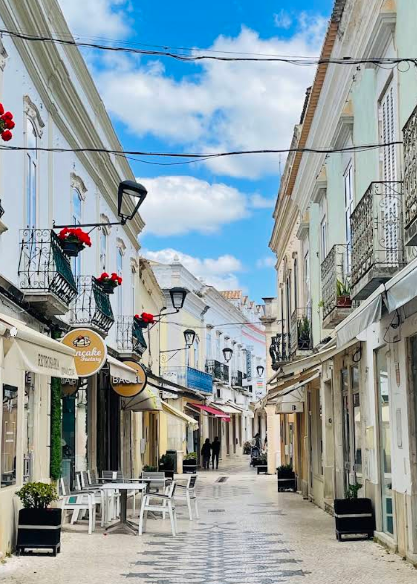 A narrow European-style street lined with white and pastel buildings under a bright blue sky with scattered clouds. Balconies with red flowers, hanging black lanterns, storefront awnings, outdoor cafe tables, and a patterned stone pavement are visible.