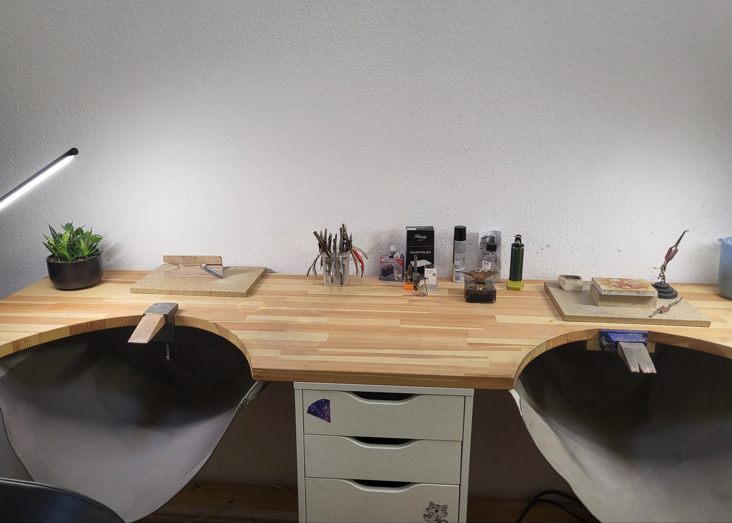 A curved wooden desk with two built-in chair seats, white drawer units underneath, and various desk accessories, tools, and a small potted plant arranged on top against a plain white wall.