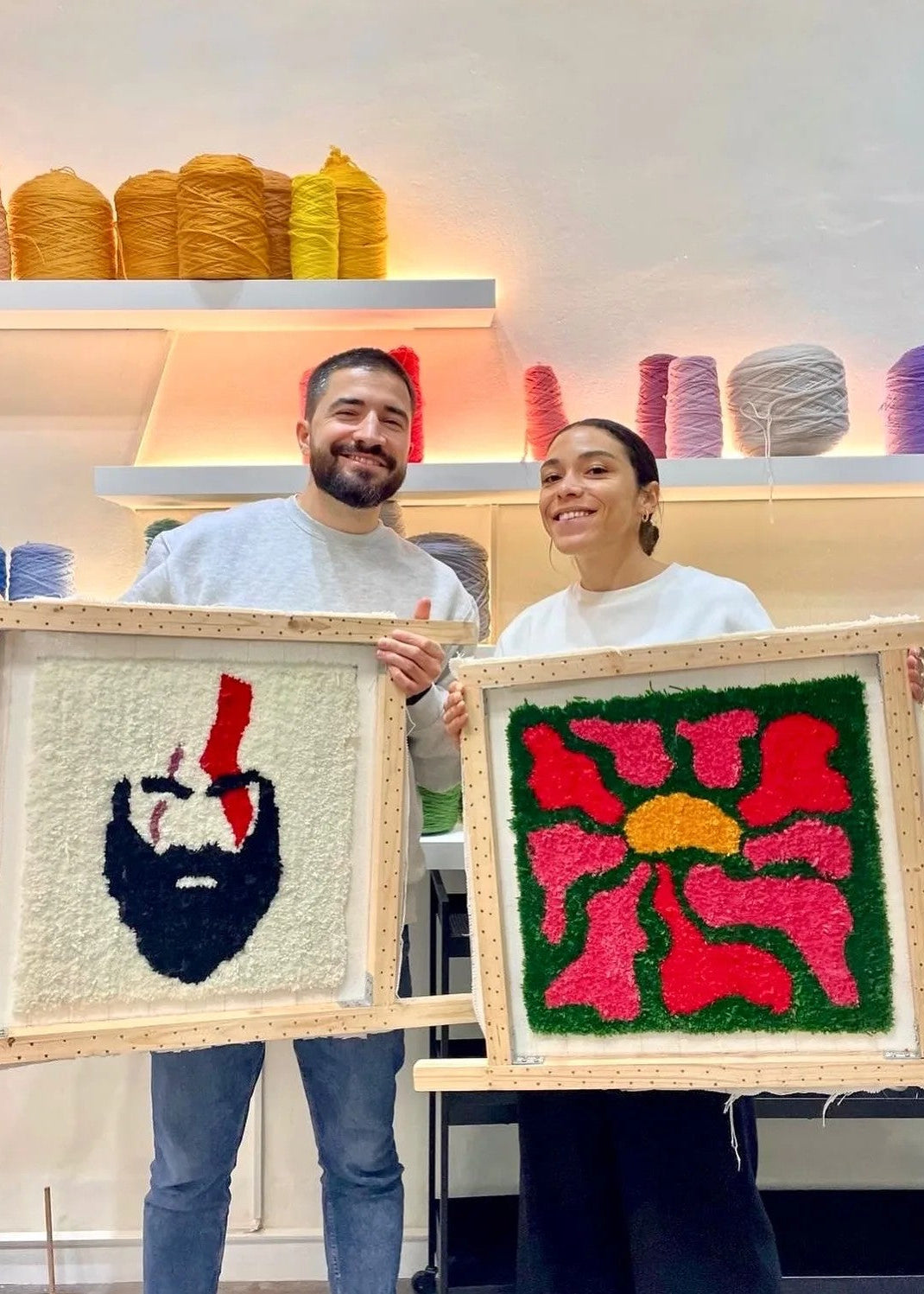 Two people holding framed woven artworks in a yarn store.