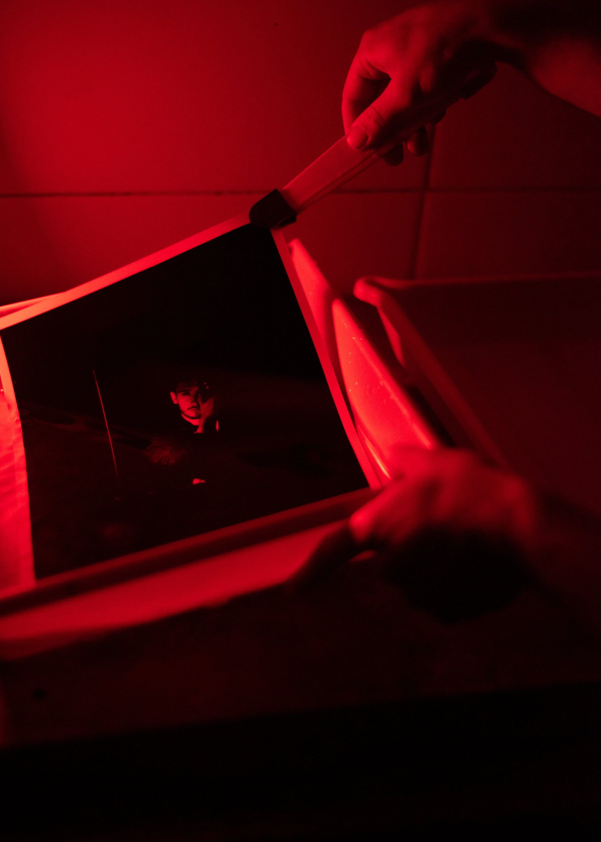 A person's hands holding a dark rectangular card or print under red light, with a tray or container nearby in a dim, monochrome red environment.