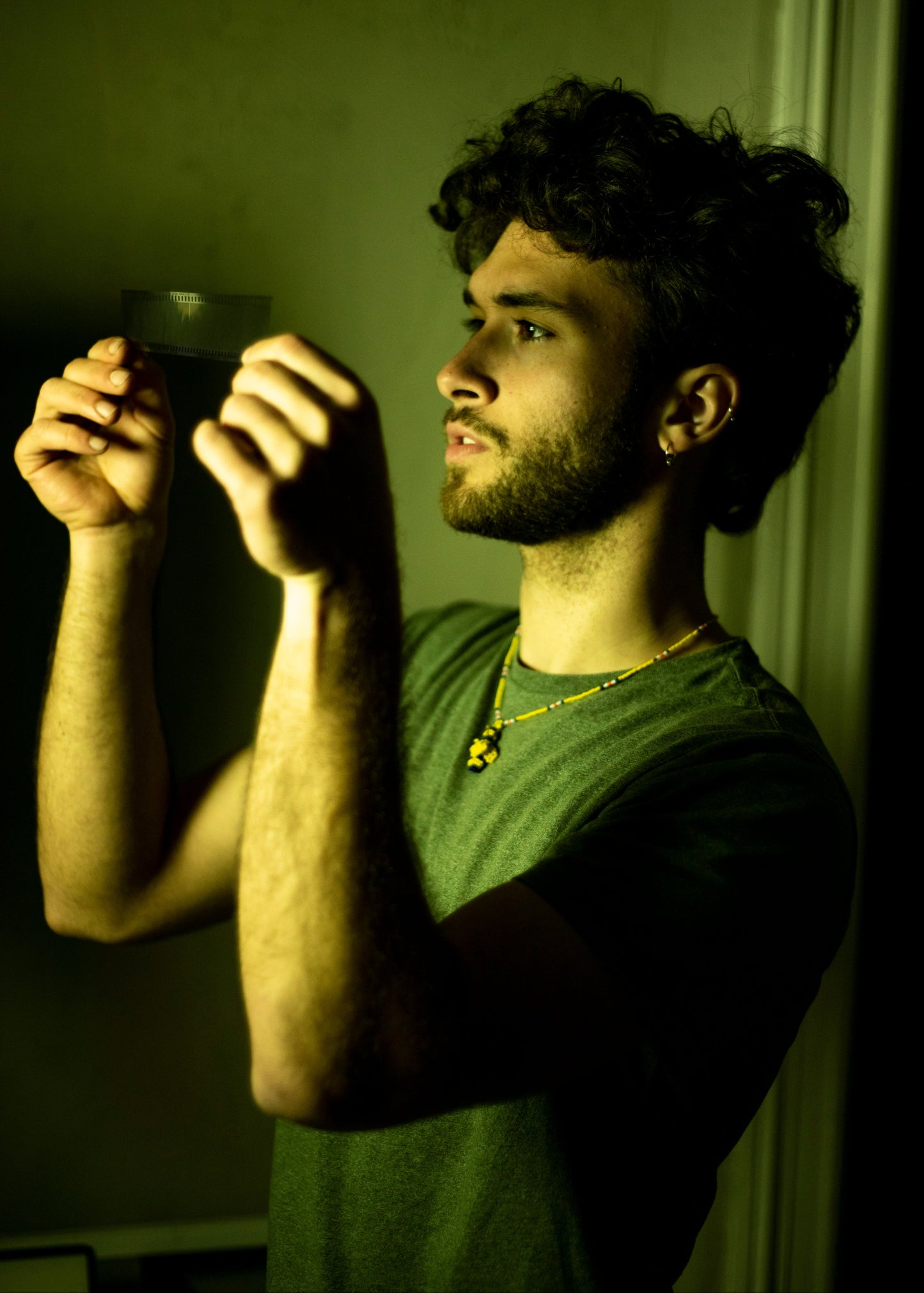 A man with curly dark hair and a beard, wearing a green t-shirt and a gold necklace, holding a small dark rectangular object up in front of his face in a dimly lit, yellow-green ambient setting.