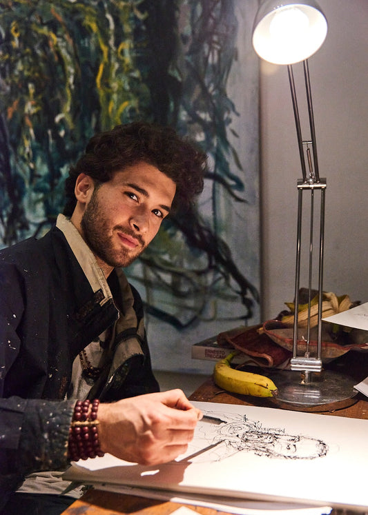 A man sits at a wooden desk under a bright adjustable lamp, sketching on paper. He is wearing a dark jacket over a patterned shirt and has a beaded bracelet on his wrist.