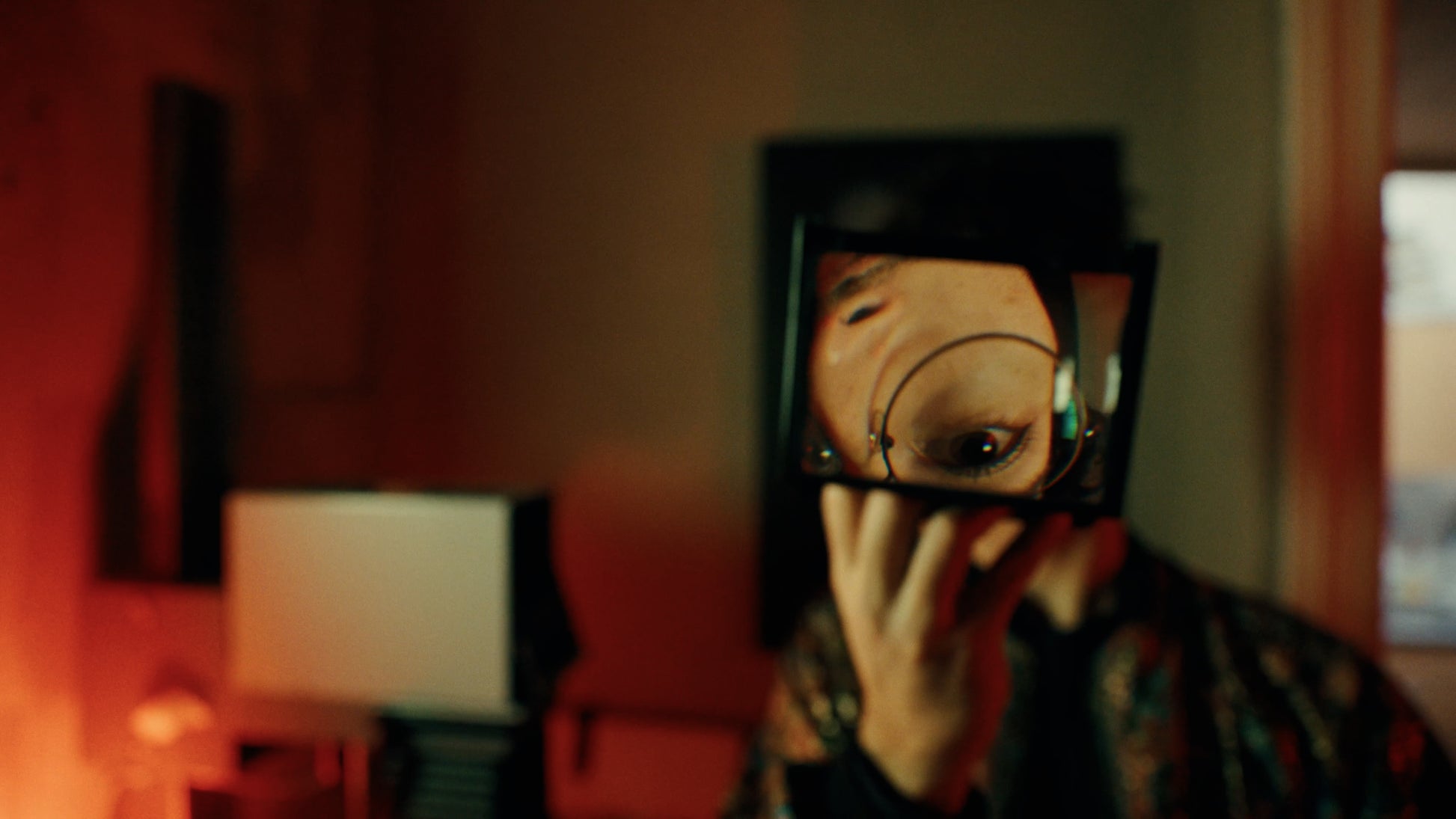 A person in a dark patterned shirt holds a small square mirror in front of their face, showing their eye and hand in a dimly lit, warm-toned room.
