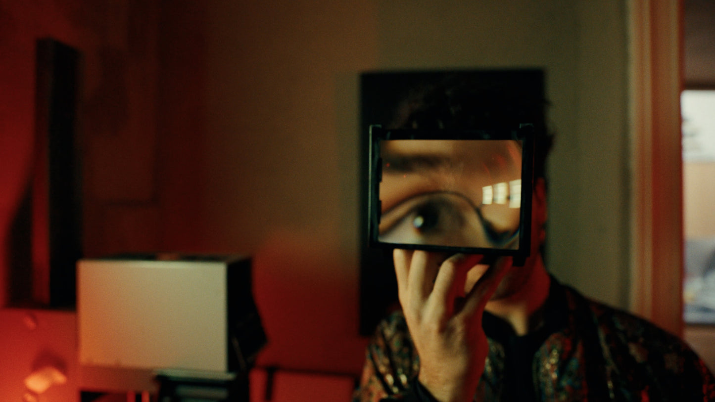 A person in a dimly lit room holds a small rectangular mirror or screen in front of their face, showing a close-up of their eye on the reflective surface.