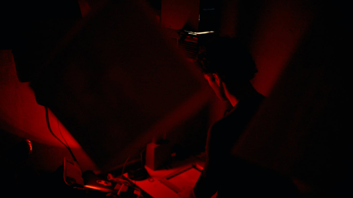 A person in a dark room illuminated by intense red lighting, viewed from behind as they wear what appears to be a headset or mask. The scene is heavily shadowed and low-light, with red casting over the surroundings.