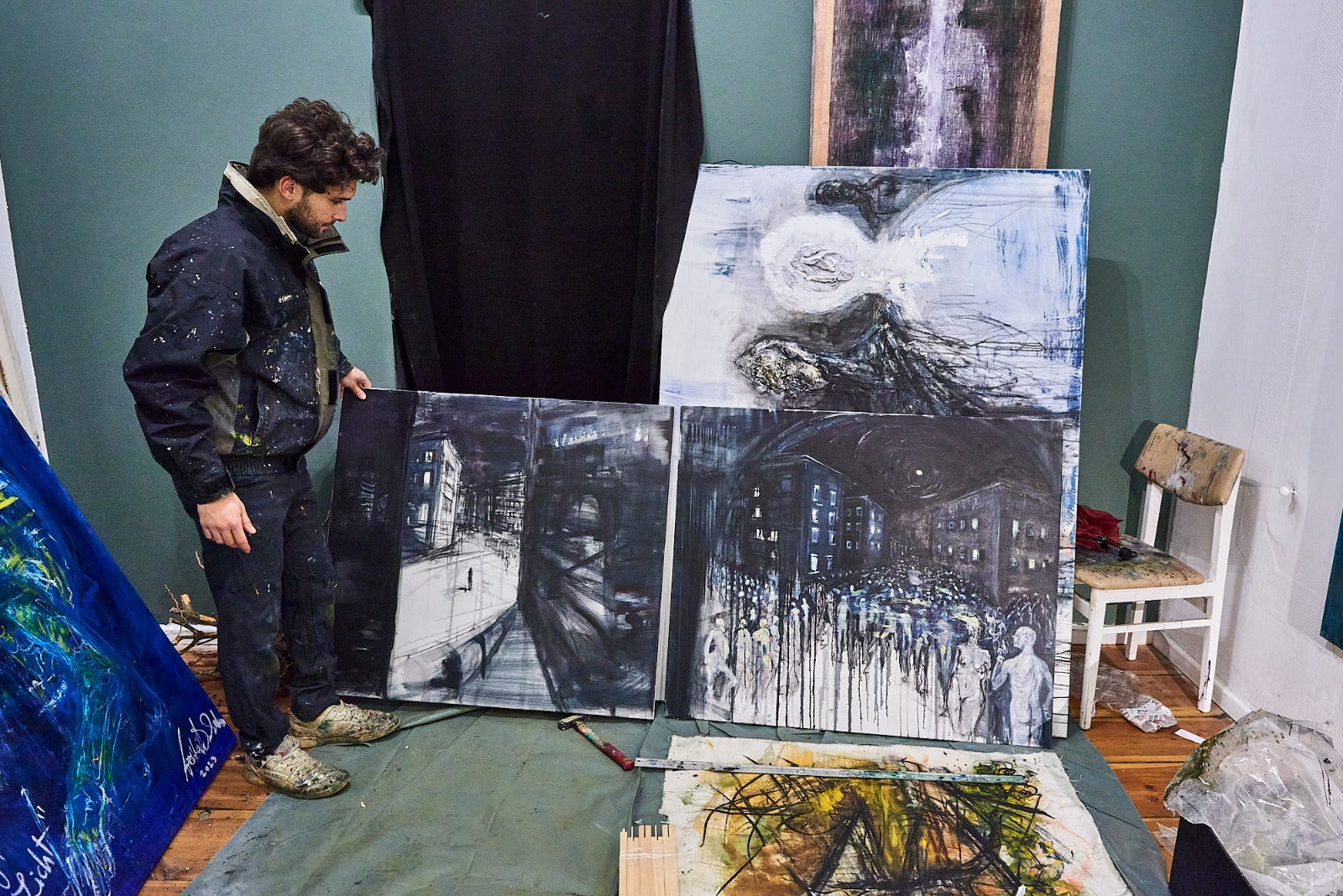 An artist in a dark jacket and jeans stands beside several large black-and-white painted canvases in an art studio, with additional paintings and a chair visible in the room.