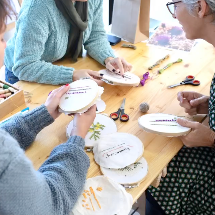 Three people seated around a wooden table participating in a craft activity, holding embroidery hoops with white fabric and embroidered floral designs. Scissors, thread, and other crafting supplies are scattered on the table.