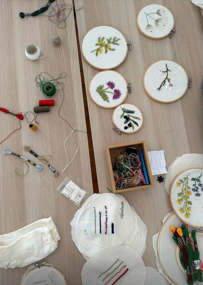 A top-down view of a wooden table covered with embroidery hoops, thread spools, embroidery floss, pins, and finished floral and botanical embroidered patches on white fabric.