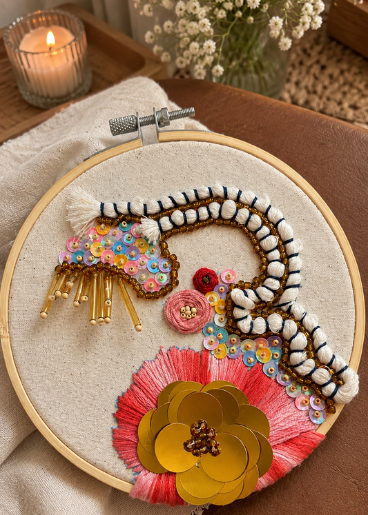 A round embroidery hoop holding a decorative textile artwork with white yarn, multicolored sequins, gold beads, pink thread, and a large gold sequin flower, photographed on a brown leather surface with a lit candle and flowers in the background.