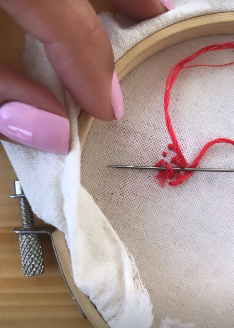 A close-up of a hand with pink manicured nails holding a wooden embroidery hoop with cream fabric, demonstrating how to tie a knot with a red thread and needle.