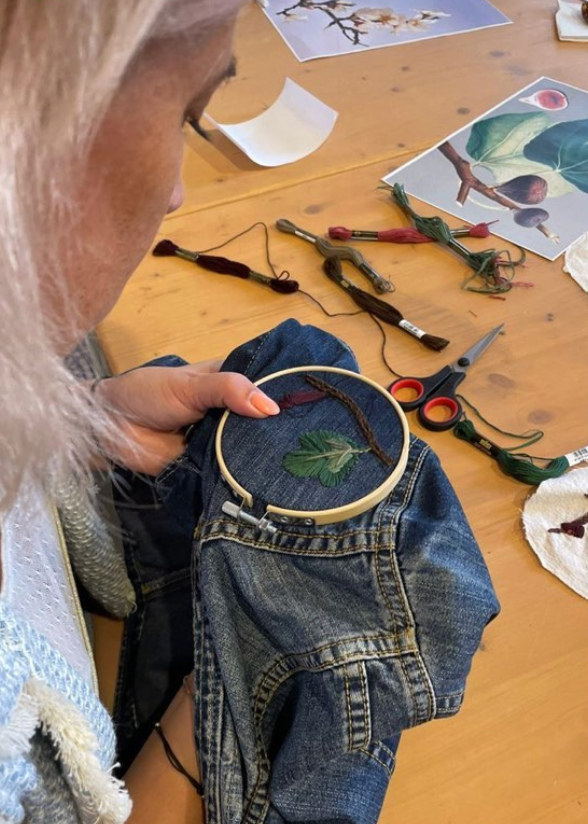 A close-up of a person holding a denim jacket while working on an embroidery project with a wooden hoop containing green leaves and brown thread, set on a wooden table surrounded by craft supplies.