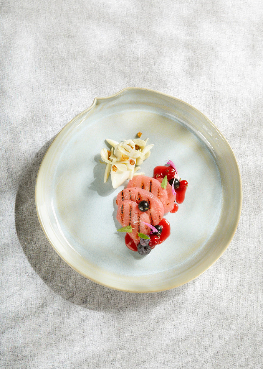 An overhead shot of a beautifully plated gourmet dish on a textured light beige linen tablecloth. The dish features grilled salmon with blackened grill marks, served with a red berry sauce, edible flowers, and a small garnish of toasted nuts on a light blue-gray ceramic plate with a gold rim.