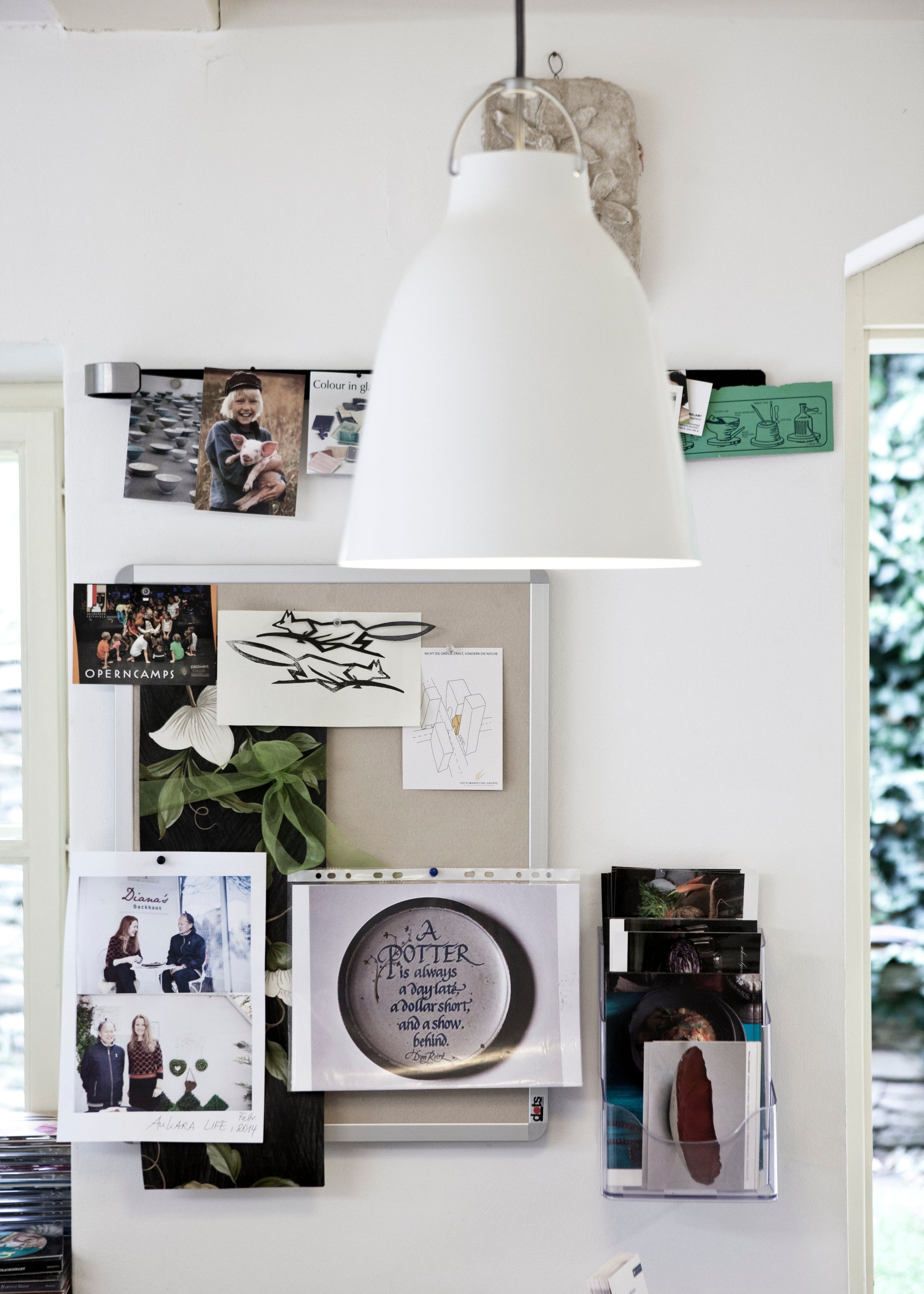 A white dome-shaped pendant light hangs in front of a white wall decorated with framed photos, a cork board with sketches and notes, a green plant, and a small clear acrylic photo holder containing a brown shoe.