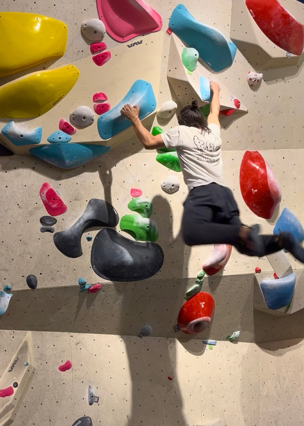 A man climbing an indoor rock wall, reaching with both hands and wearing a white t-shirt and dark shorts. The wall features numerous colorful climbing holds in blue, red, yellow, pink, green, and gray against a speckled beige background.