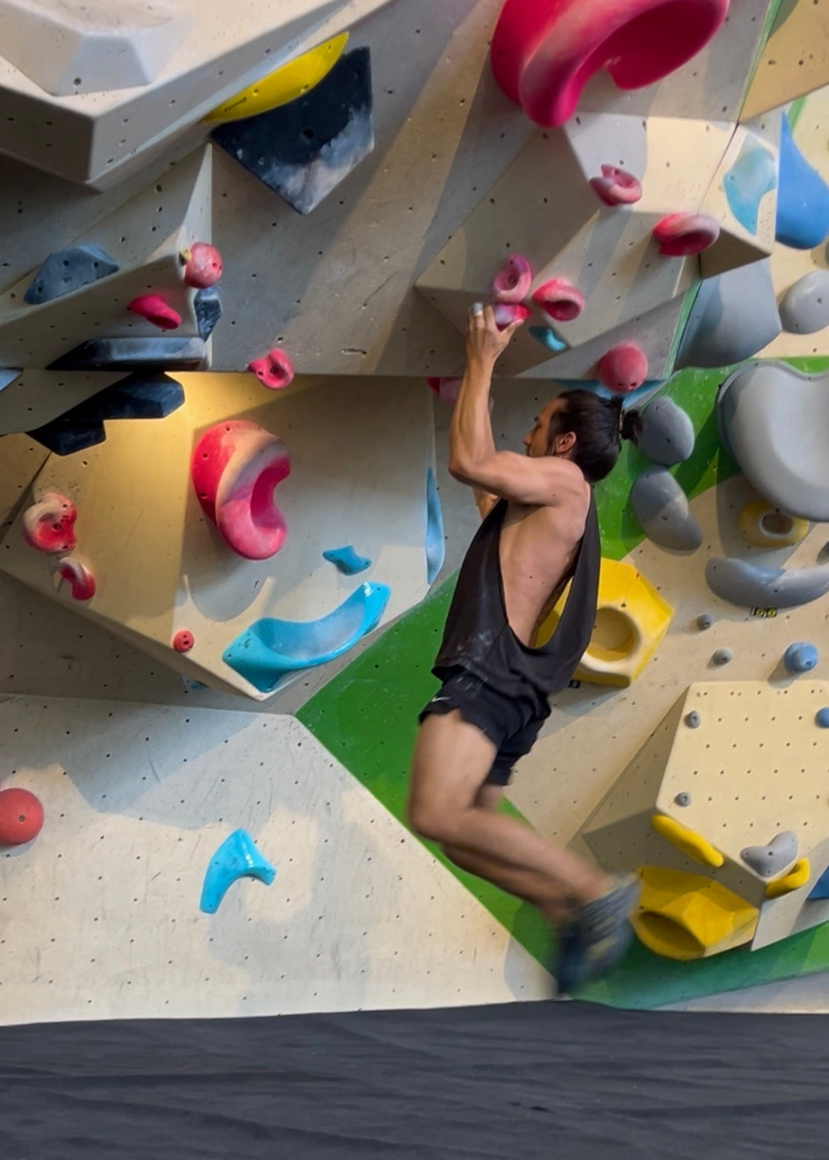 A person climbing an indoor rock wall, wearing a sleeveless black top and dark shorts, with colorful climbing holds and a textured wall background.