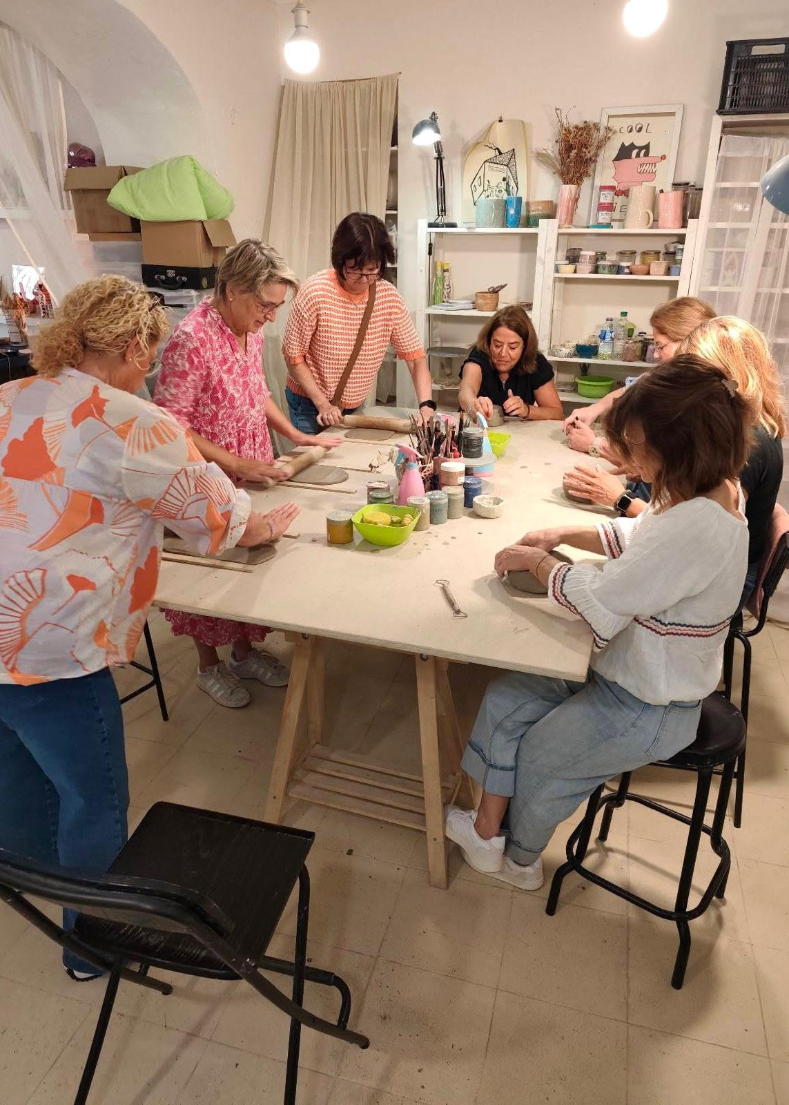 A group of women gathered around a wooden table in a bright, creative studio, working on crafts or painting. The room features white walls, shelves filled with art supplies, and hanging light bulbs.