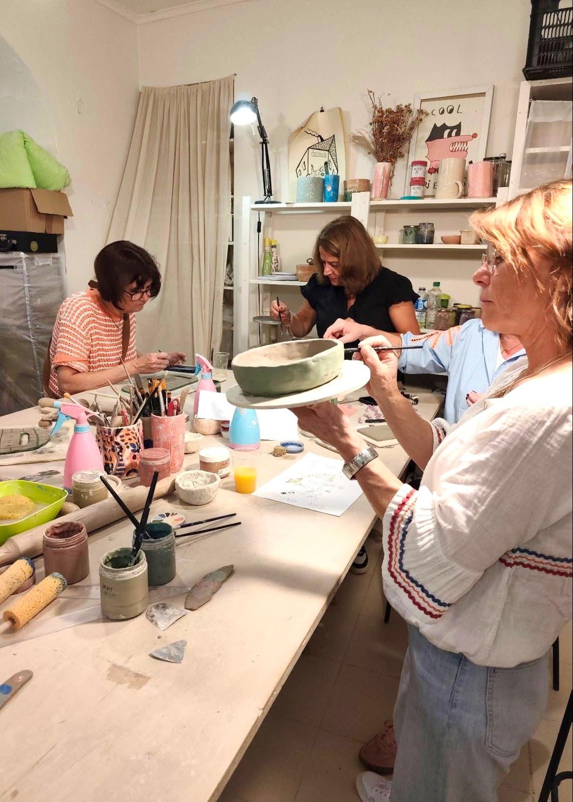 A pottery workshop scene with several women gathered around a table, working with clay and pottery tools. One woman in the foreground holds a green ceramic bowl on a white turntable.