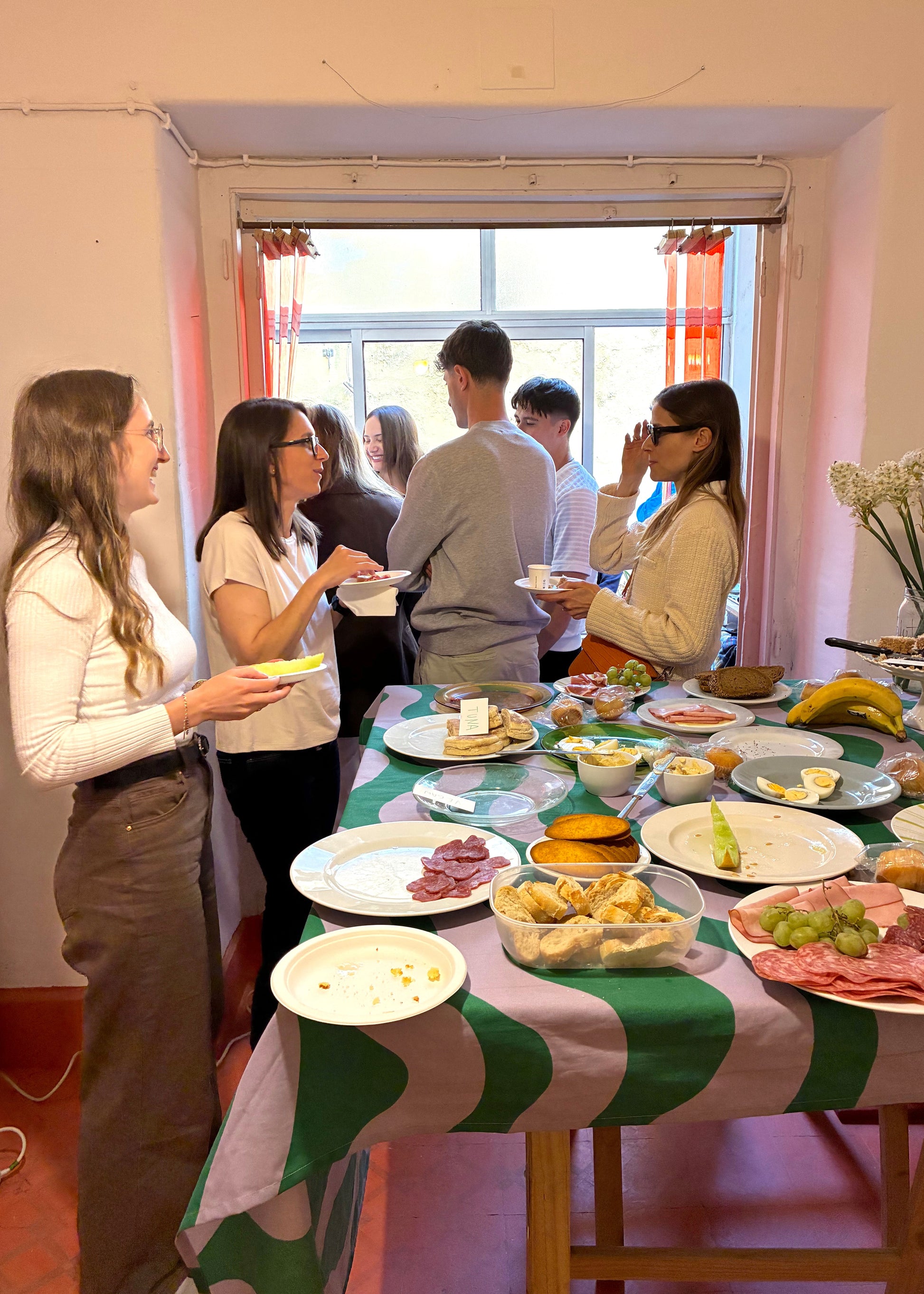 People gathered around a table with food in a room with large windows and curtains.