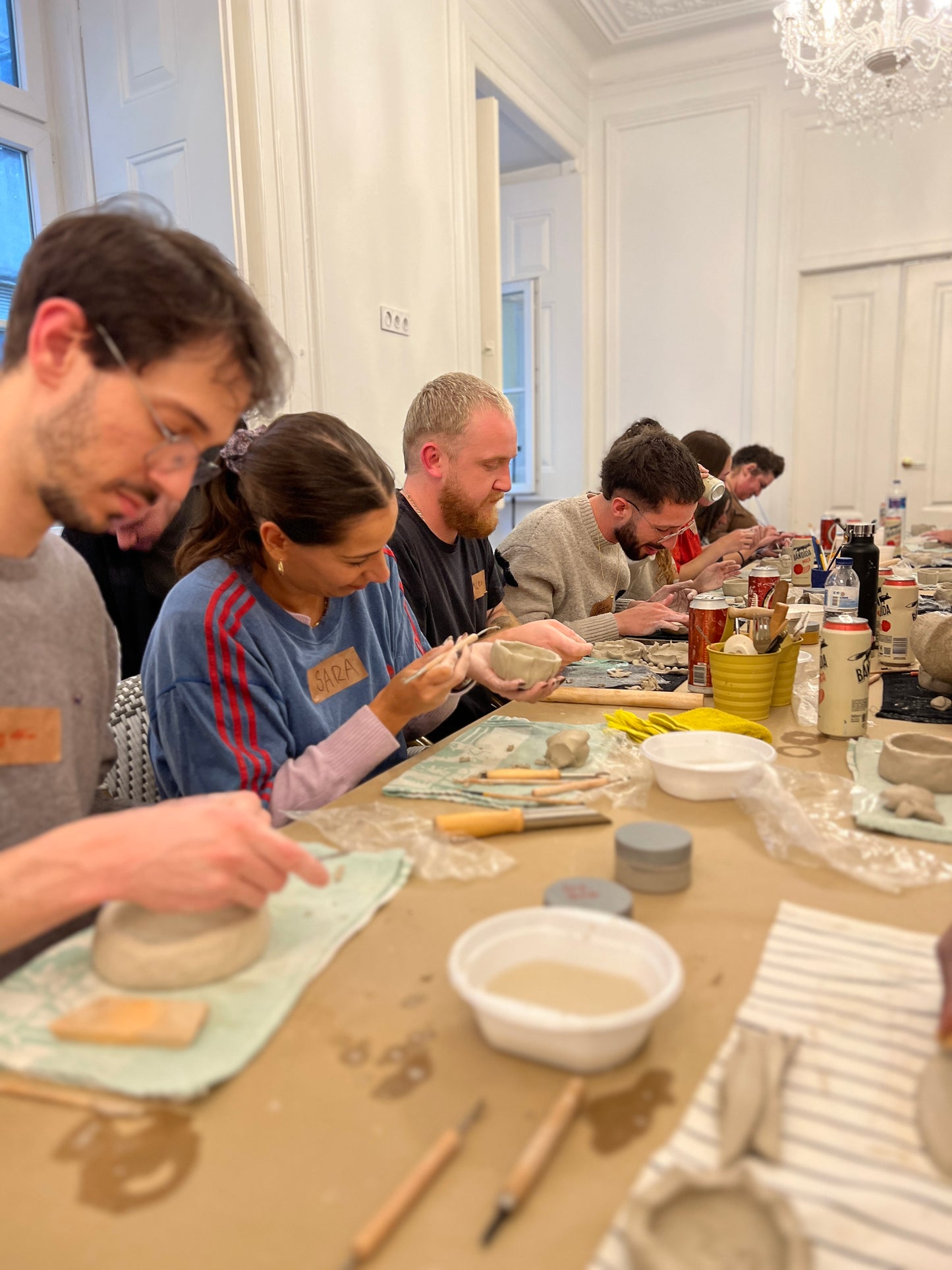 Group of people working on pottery at a table in a room with white walls and a chandelier.
