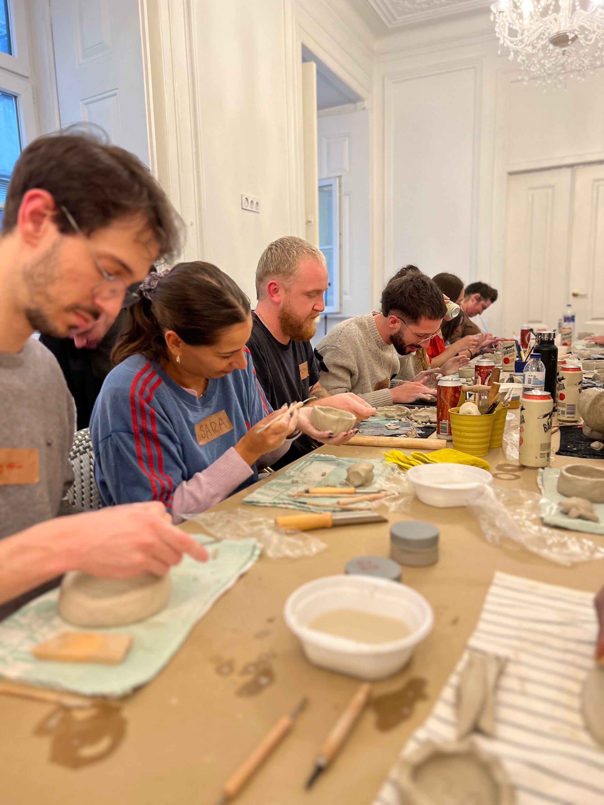 Group of people working on pottery at a table in a room with white walls and a chandelier.