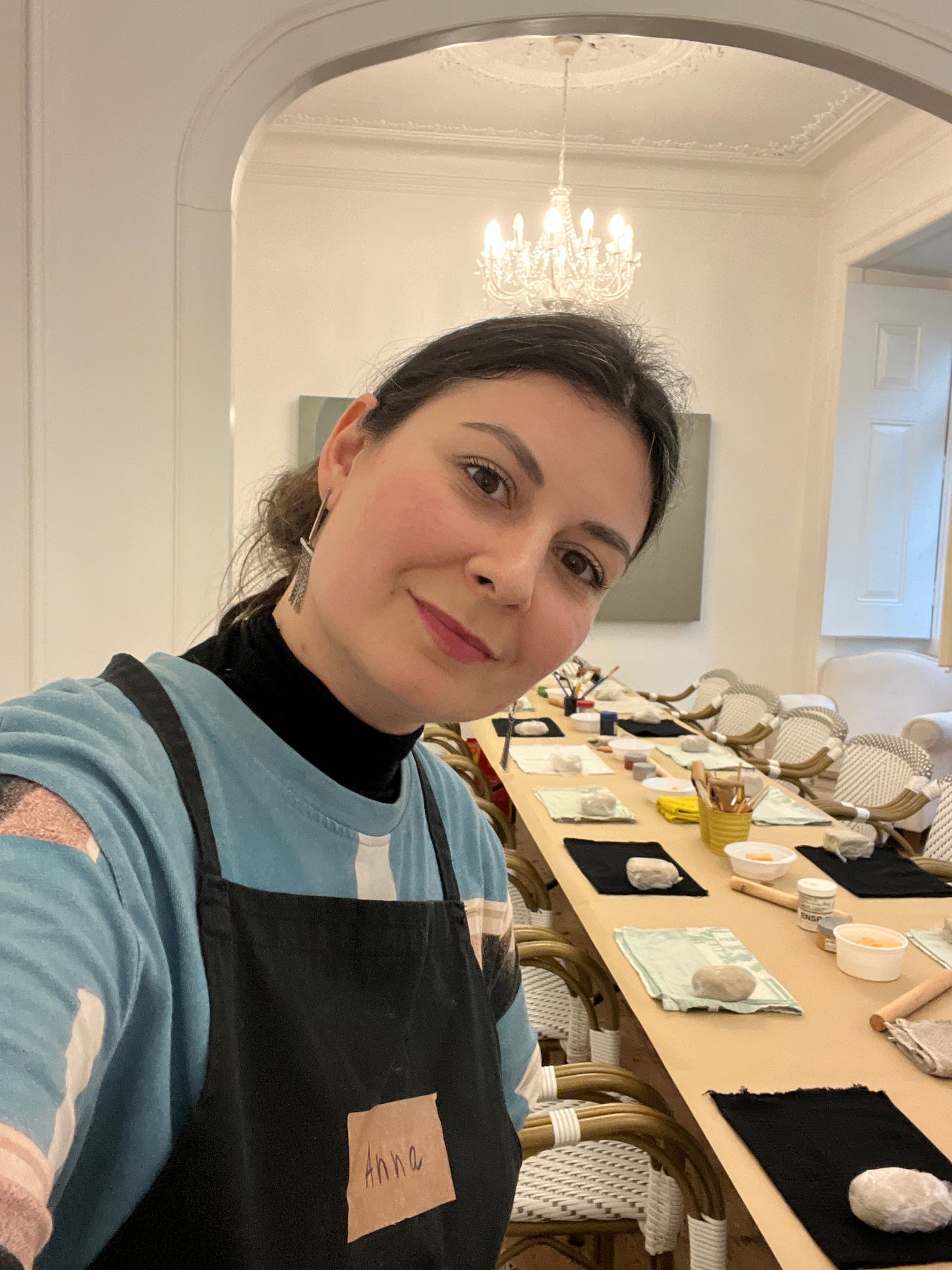 Woman wearing a blue sweater with a black apron in a room with decorative items on shelves.