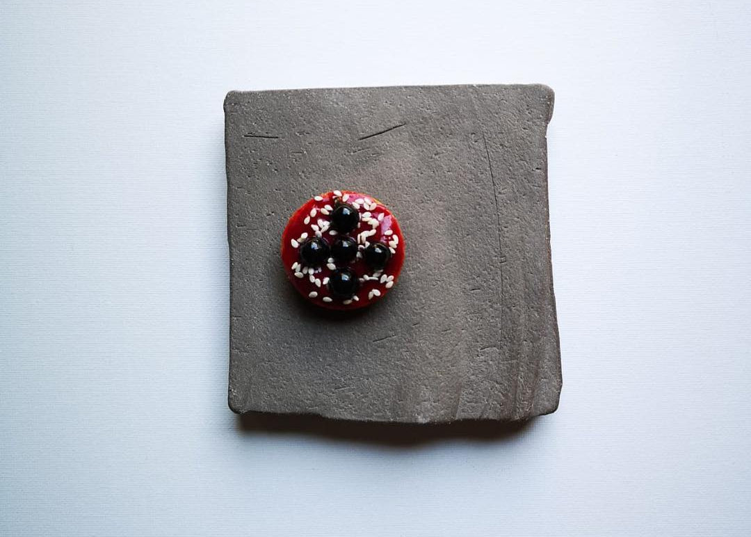 A small round red button with white and black speckles, placed on a square textured gray slate coaster against a light gray background.