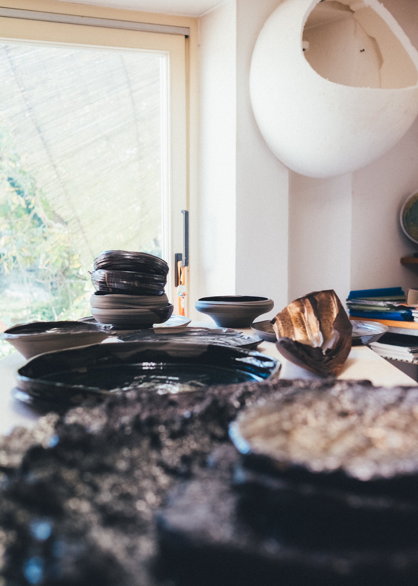A collection of handmade ceramic dishes, including a stack of dark bowls, a shallow dish, and a textured wooden spoon, arranged on a table in a bright room with a window and a white hanging pod chair in the background.