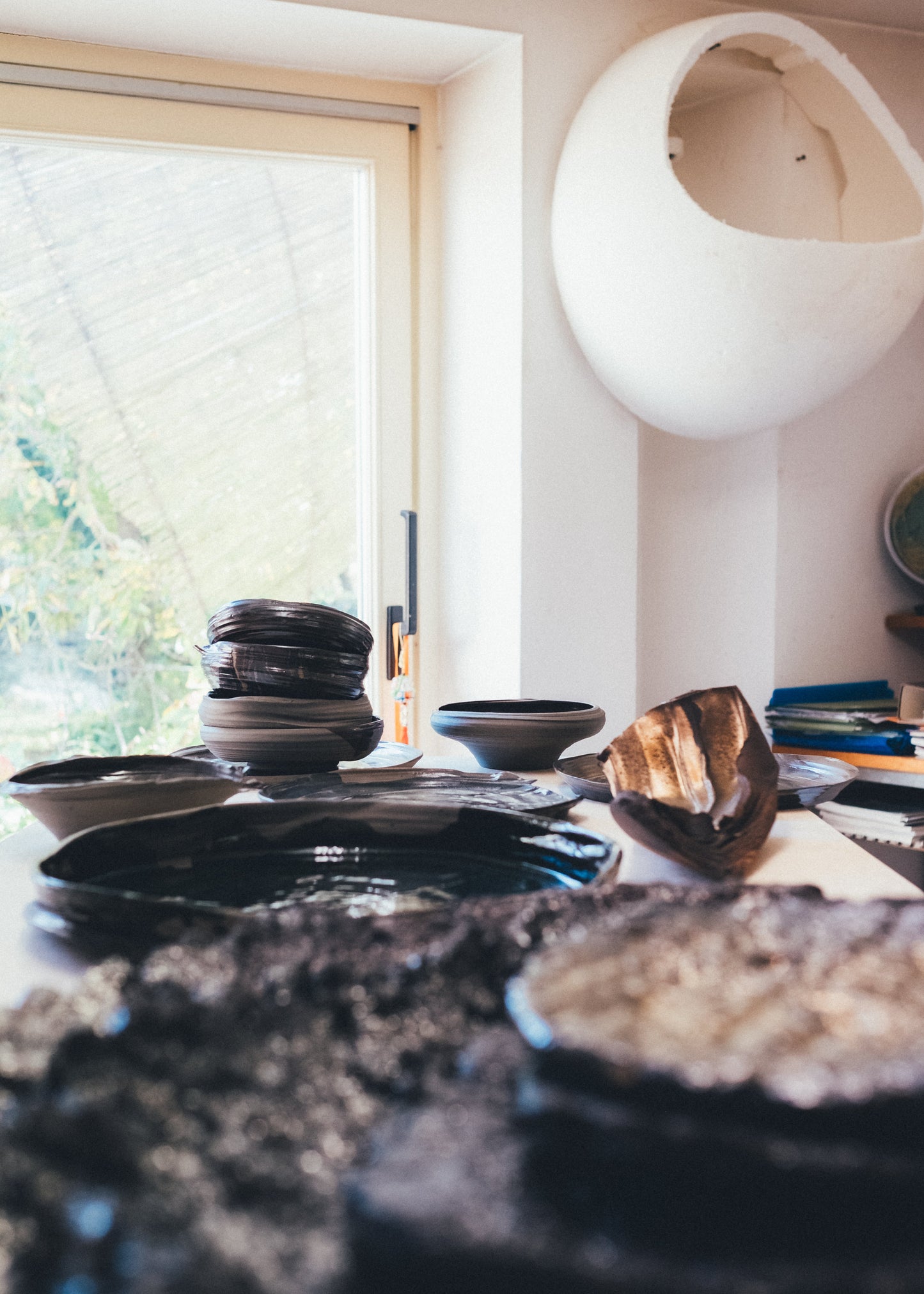 A collection of handmade ceramic dishes, including a stack of dark bowls, a shallow dish, and a textured wooden spoon, arranged on a table in a bright room with a window and a white hanging pod chair in the background.