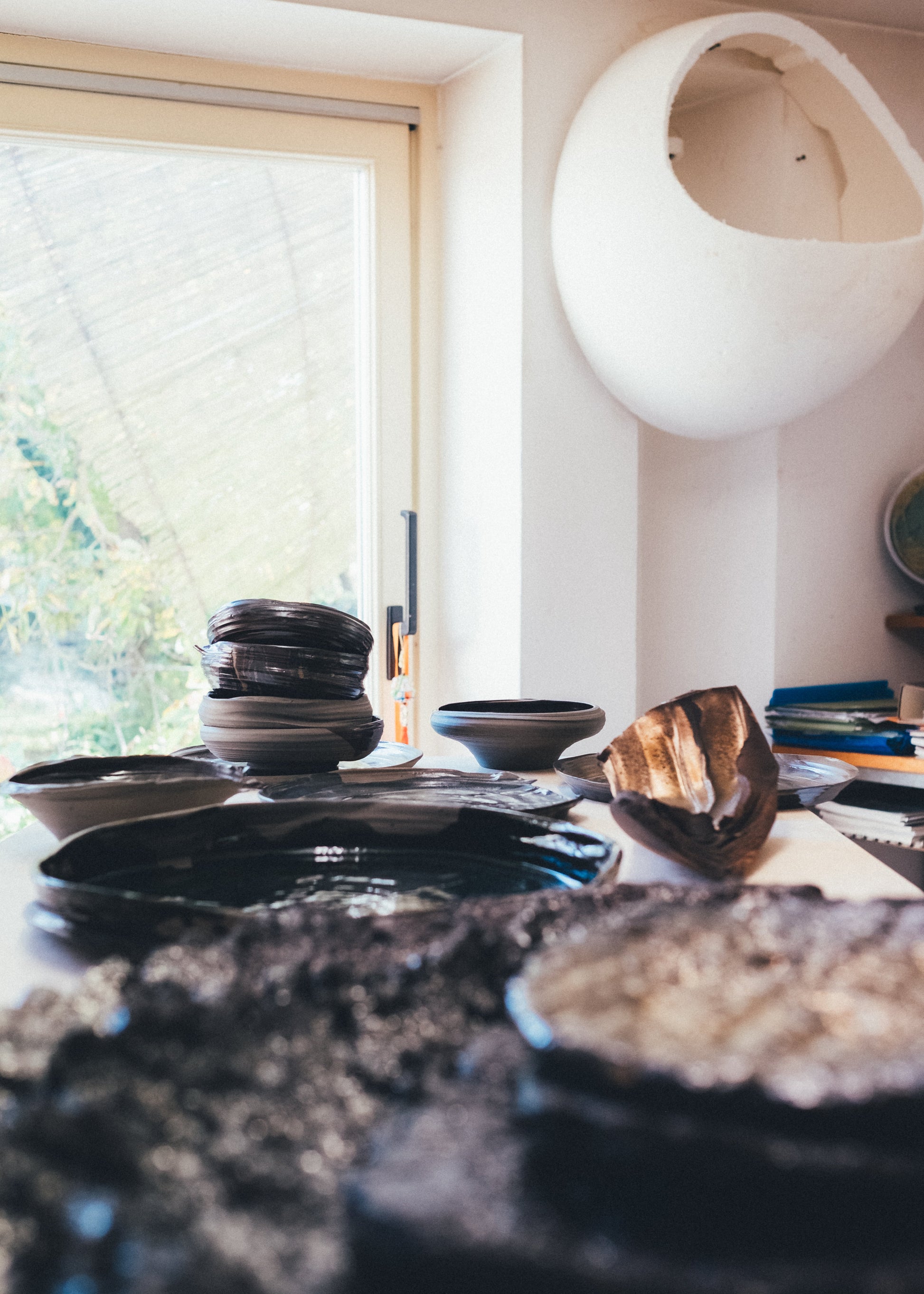 A collection of handmade ceramic dishes, including a stack of dark bowls, a shallow dish, and a textured wooden spoon, arranged on a table in a bright room with a window and a white hanging pod chair in the background.