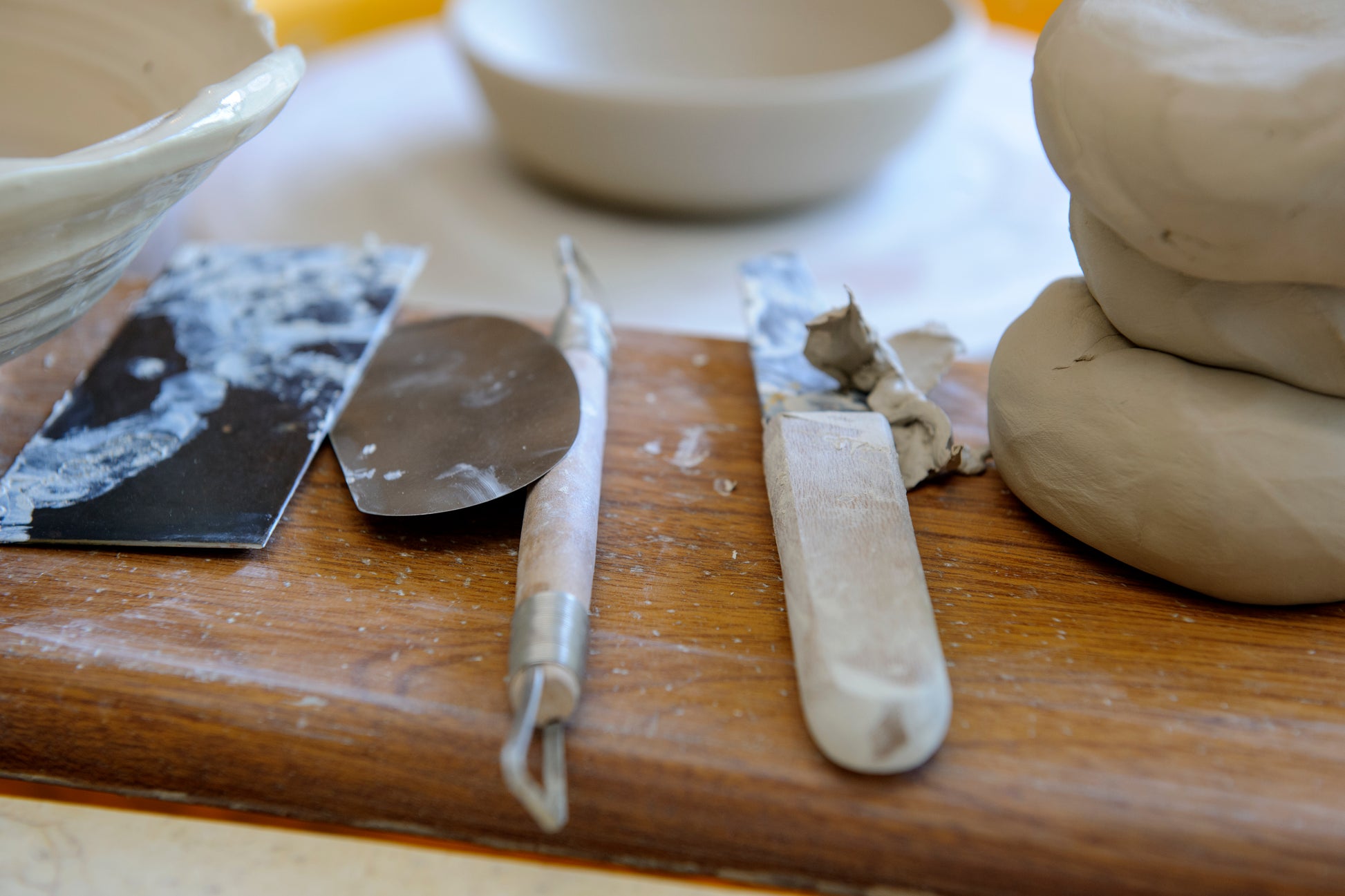 A close-up of pottery tools resting on a wooden surface beside clay dough, including a metal spatula, a sculpting knife, and stacked clay balls.