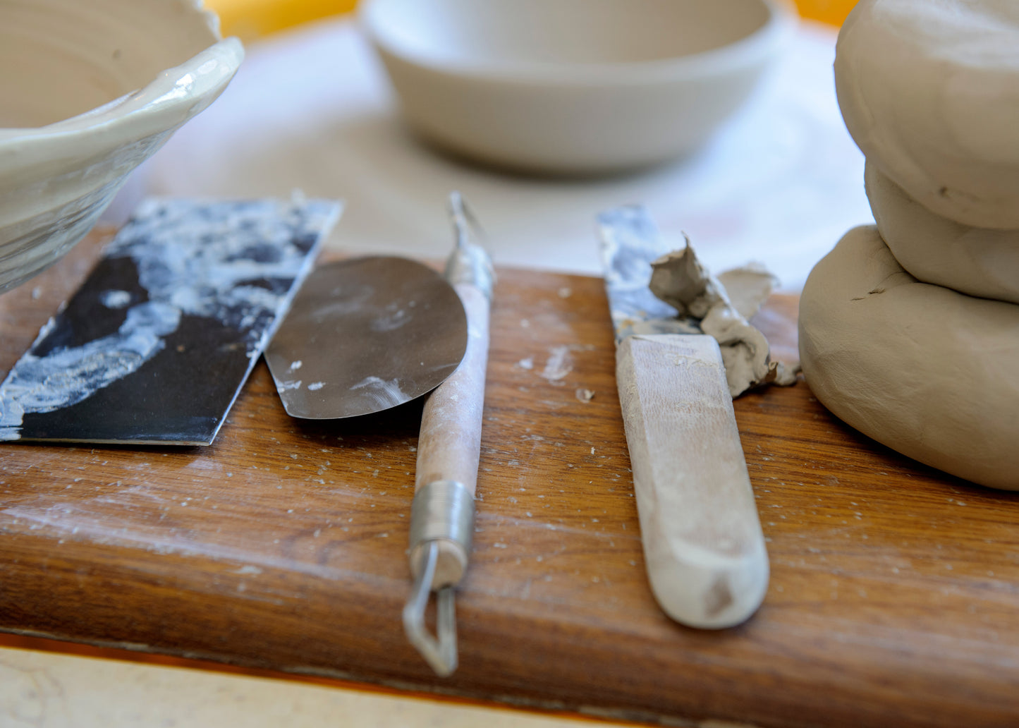 A close-up of pottery tools resting on a wooden surface beside clay dough, including a metal spatula, a sculpting knife, and stacked clay balls.