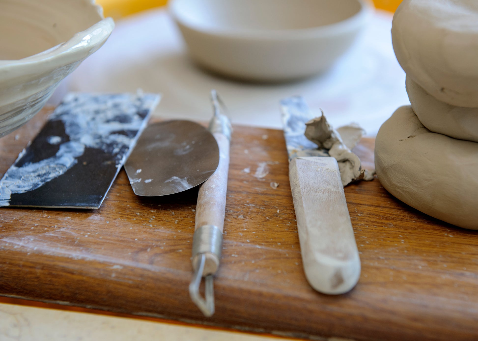 A close-up of pottery tools resting on a wooden surface beside clay dough, including a metal spatula, a sculpting knife, and stacked clay balls.