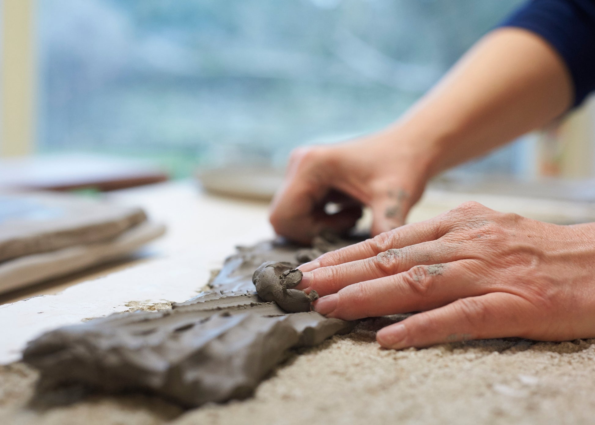 Close-up of a person's hands shaping a piece of gray clay on a sandy surface, with a blurred workshop background.