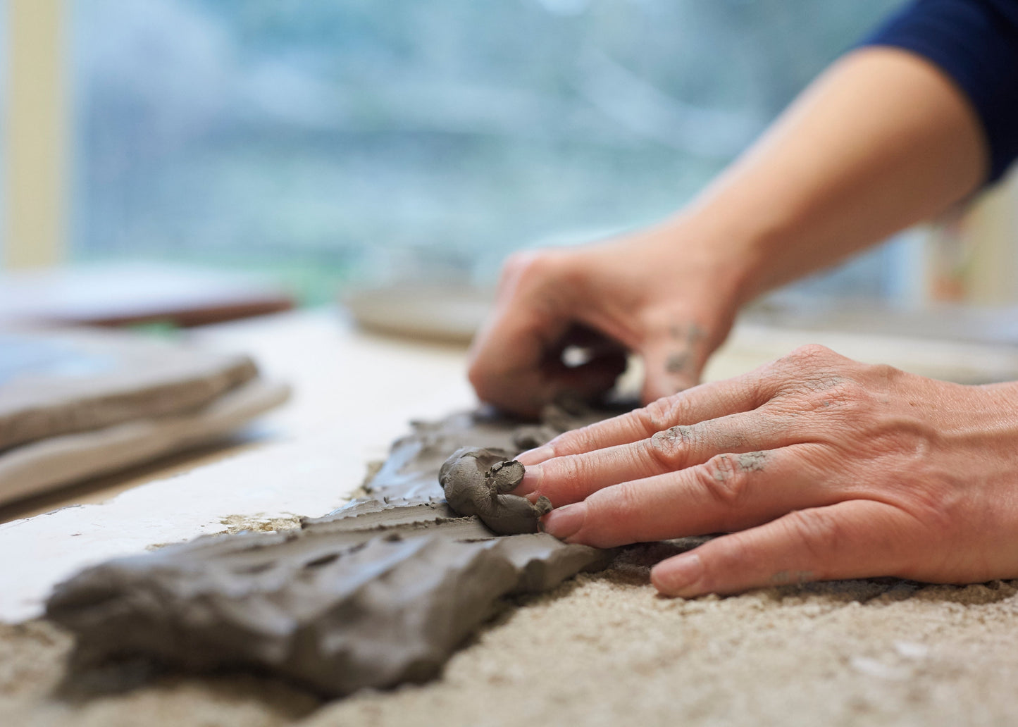 Close-up of a person's hands shaping a piece of gray clay on a sandy surface, with a blurred workshop background.