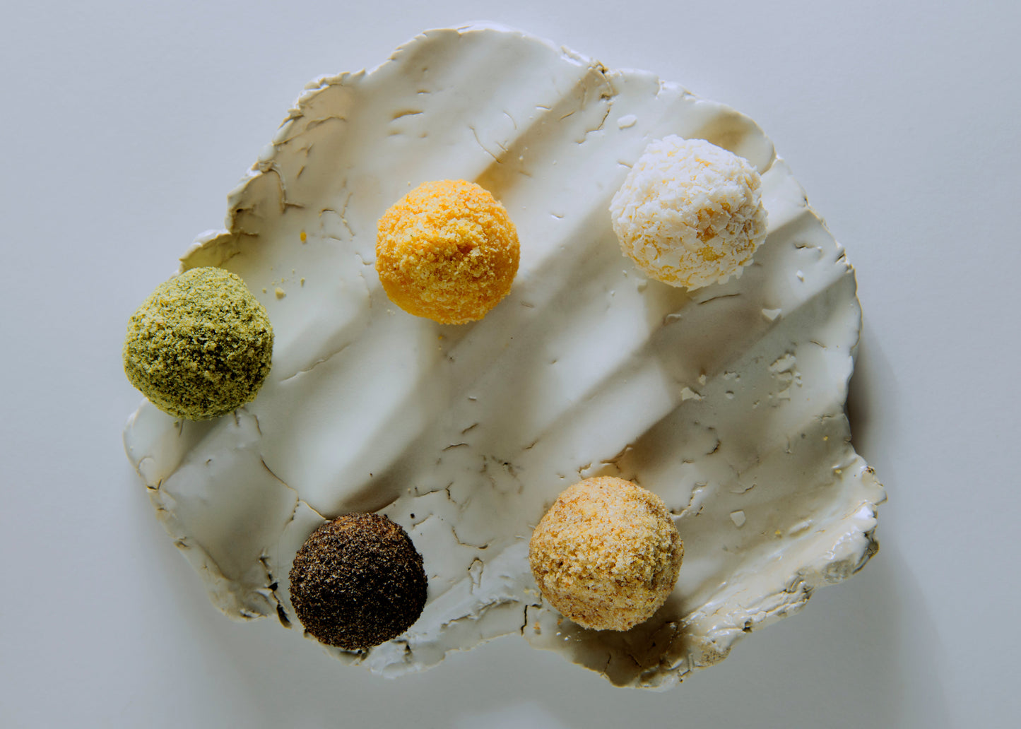 Five small spherical food items in different colors arranged on a textured off-white plate against a plain light background.