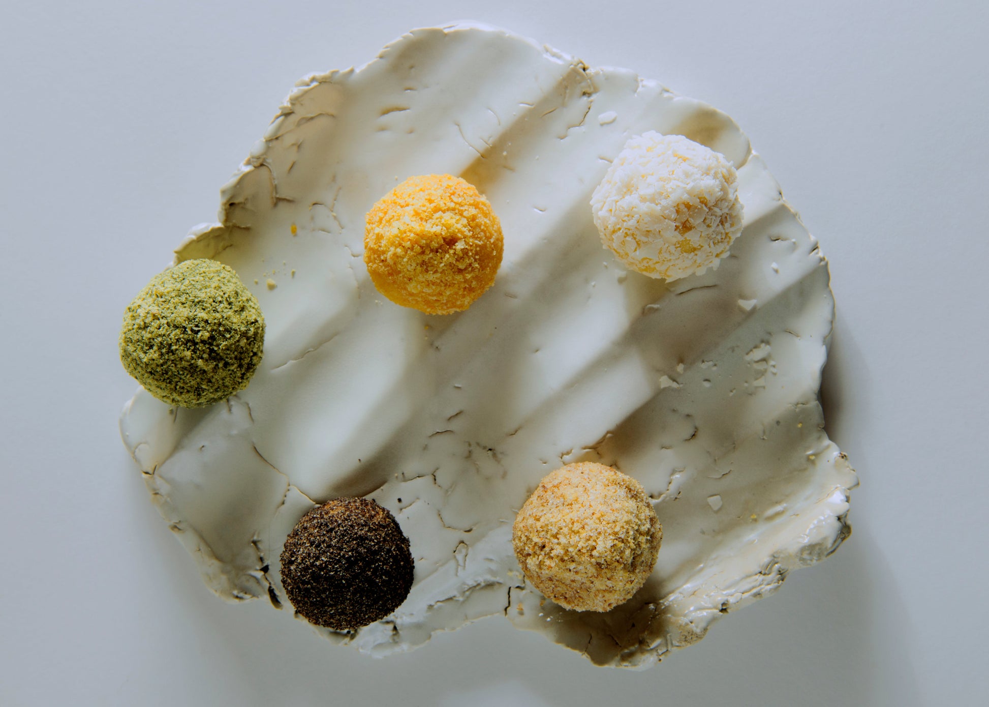 Five small spherical food items in different colors arranged on a textured off-white plate against a plain light background.