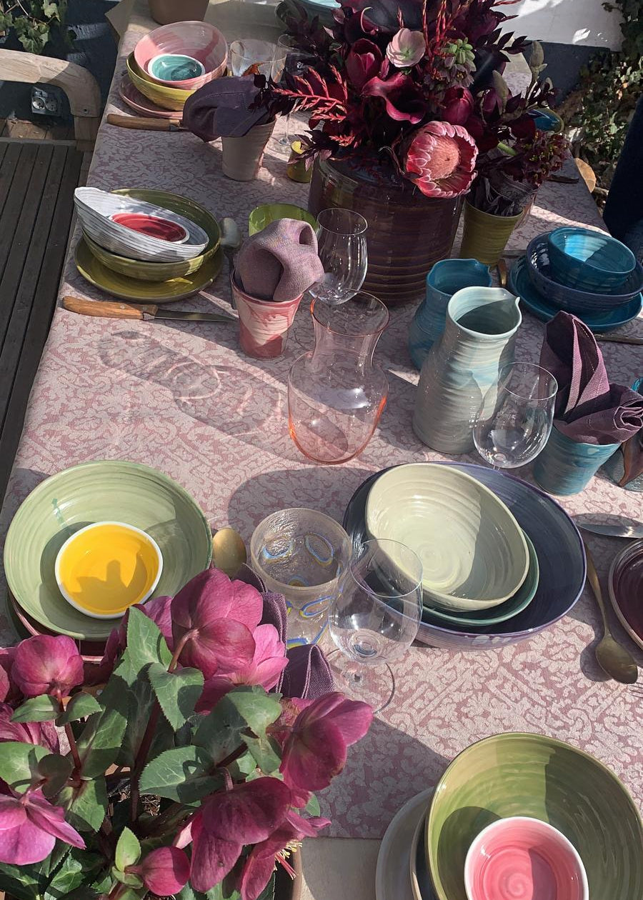 A styled outdoor dining table set with multiple pastel-colored ceramic plates, bowls, and vases, decorated with pink and burgundy flowers under bright sunlight.