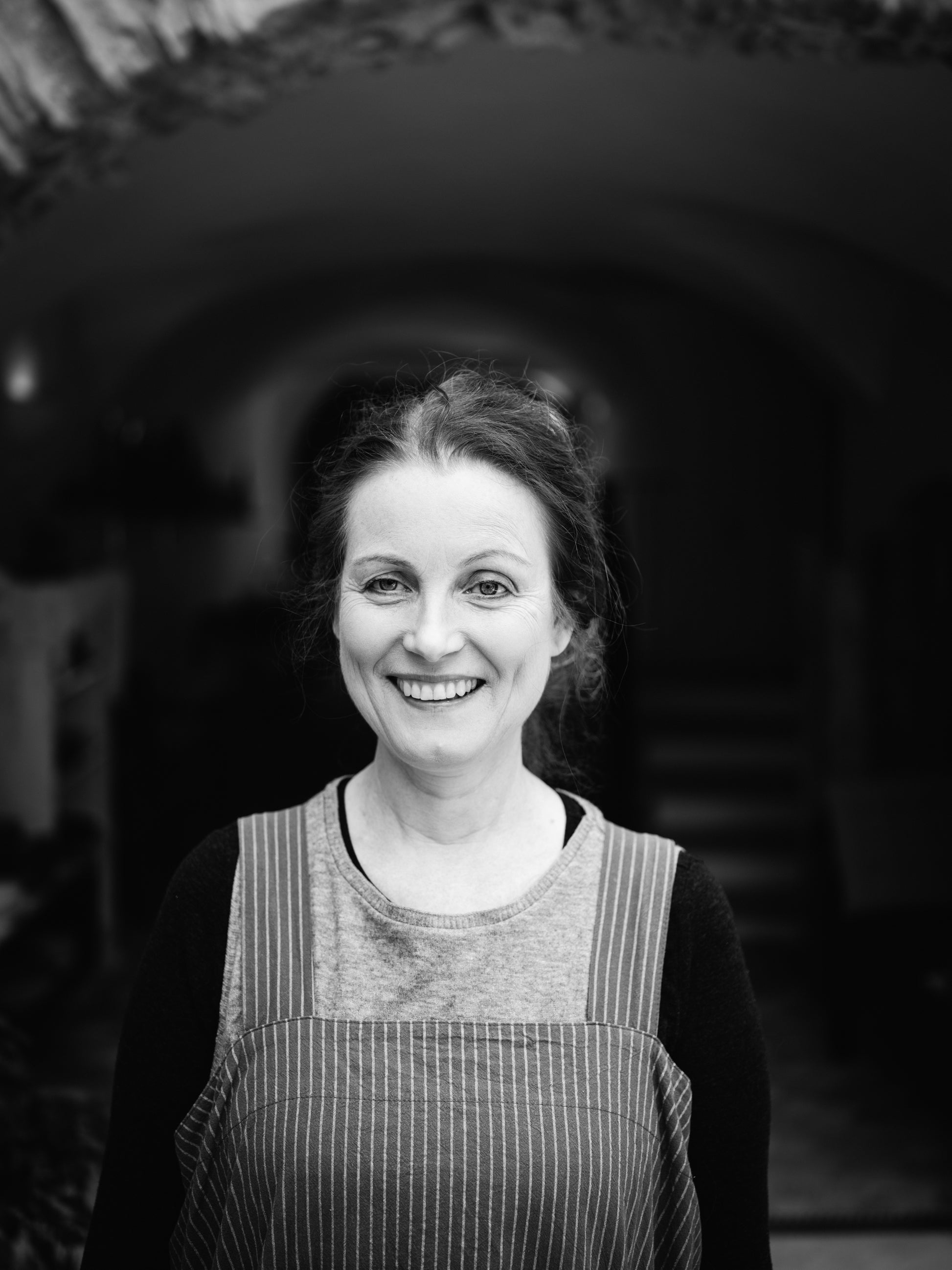 A black and white portrait of a smiling woman with her hair pulled back, wearing a striped apron over a dark long-sleeve top, standing in front of a dark arched doorway.