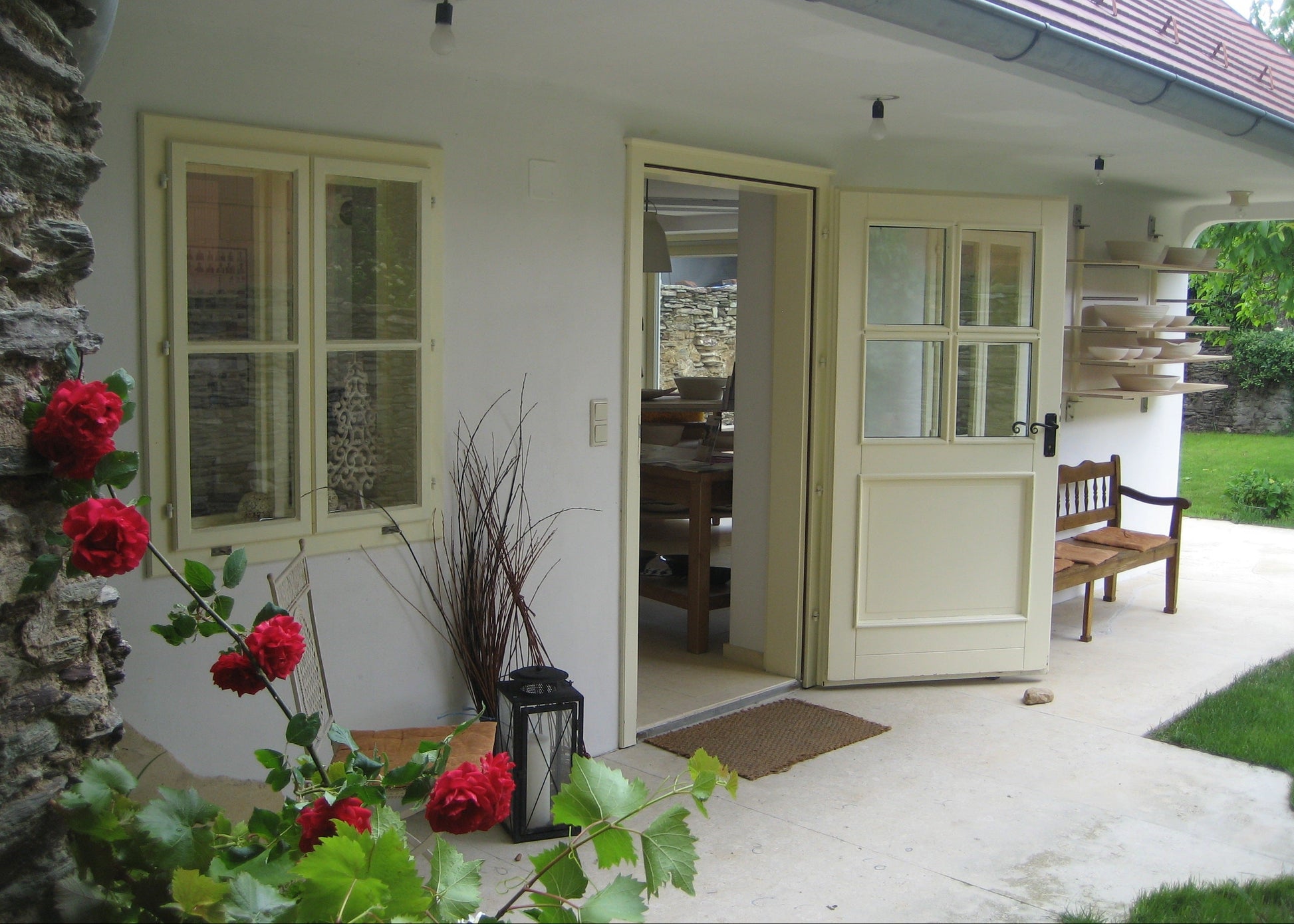 A set of cream-colored exterior double doors with glass panes and a matching window on a white stucco house, with an open doorway revealing a dining area inside and a wooden bench on the patio.