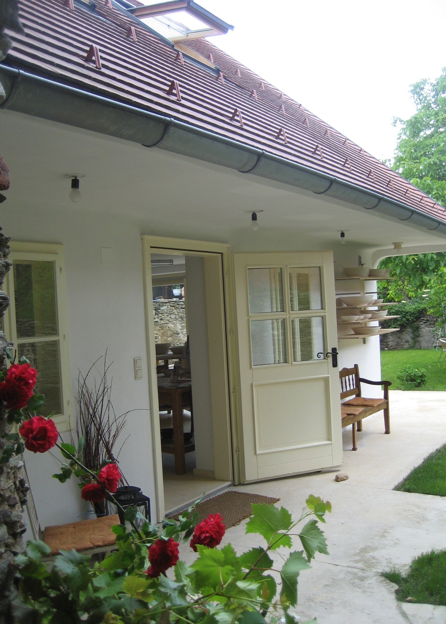 A white exterior door with six glass panes stands open on a covered patio, showing an interior dining area. The door is part of a house with a brown tiled roof, stone wall, bench, and green lawn in the background.