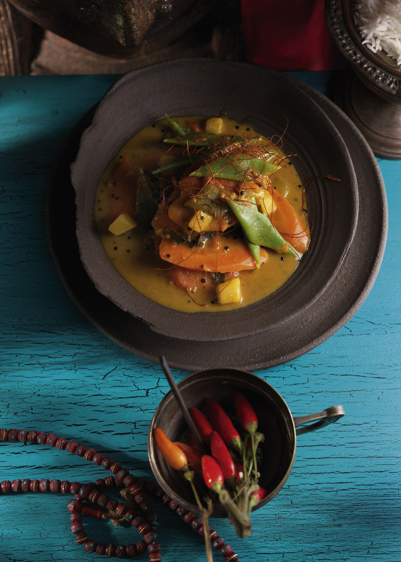 A plated curry dish with orange slices, green vegetables, and yellow sauce on a dark plate, set on a distressed turquoise wooden table with a small bowl of red chilies in the foreground.