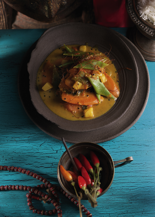 A plated curry dish with orange slices, green vegetables, and yellow sauce on a dark plate, set on a distressed turquoise wooden table with a small bowl of red chilies in the foreground.