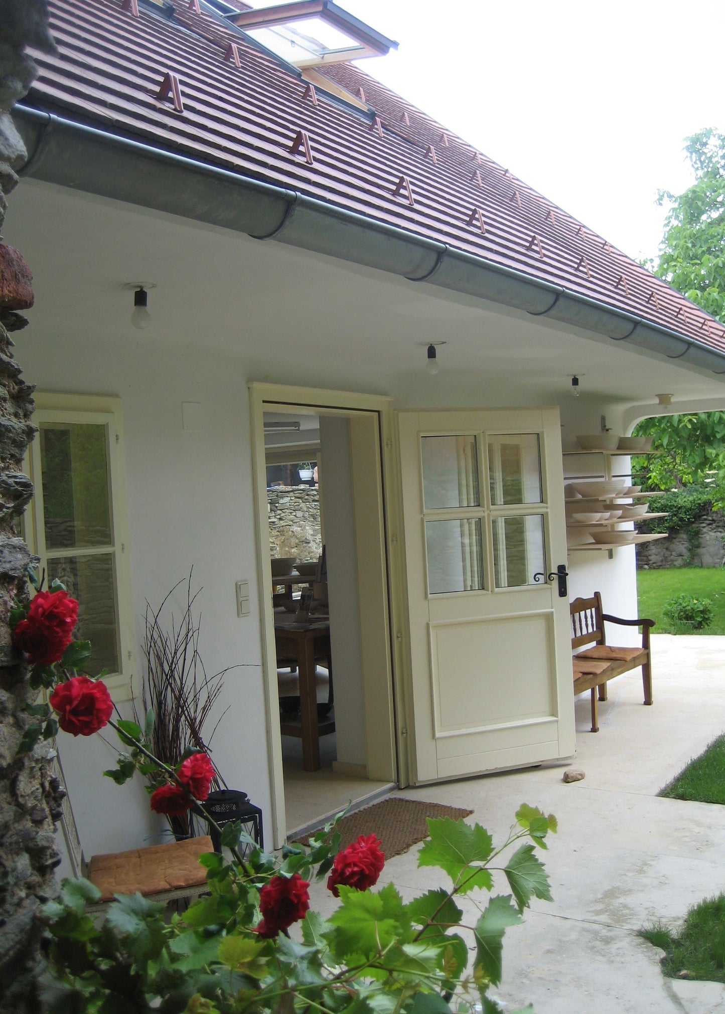 A white exterior door with six glass panes stands open on a covered patio, showing an interior dining area. The door is part of a house with a brown tiled roof, stone wall, bench, and green lawn in the background.