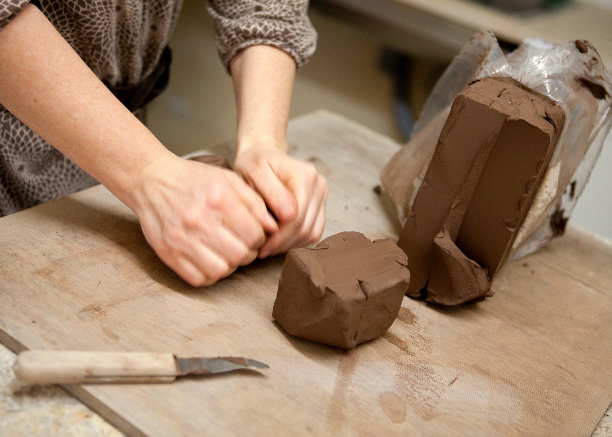 A close-up of a person's hands kneading brown clay on a wooden workbench, with a wrapped block of clay and a small sculpting knife nearby.