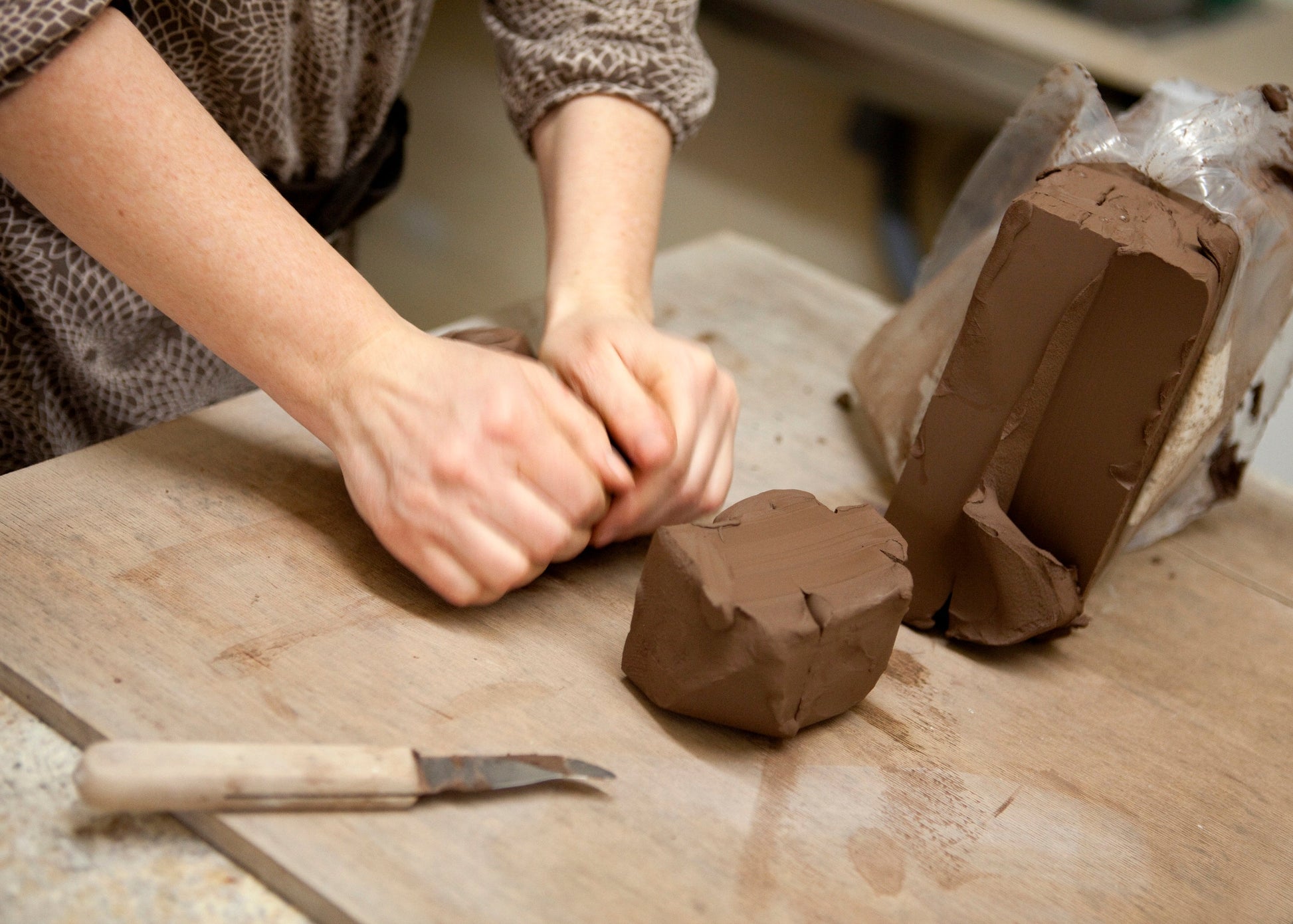 A close-up of a person's hands kneading brown clay on a wooden workbench, with a wrapped block of clay and a small sculpting knife nearby.