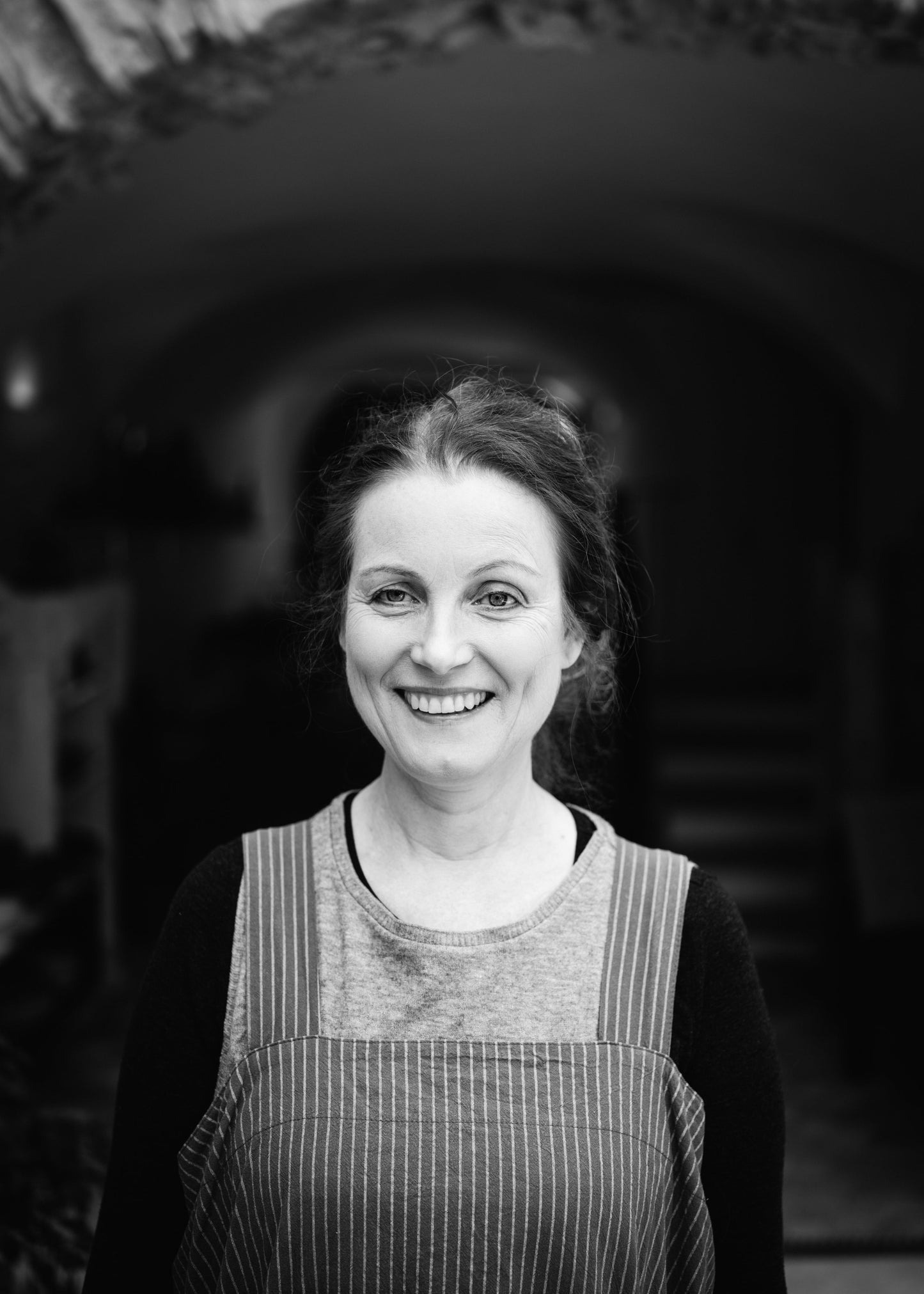 A black and white portrait of a smiling woman with her hair pulled back, wearing a striped apron over a dark long-sleeve top, standing in front of a dark arched doorway.