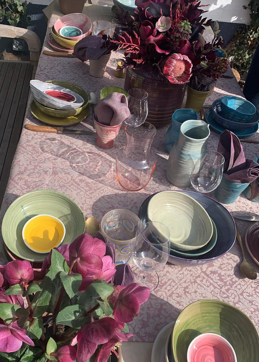 A styled outdoor dining table set with multiple pastel-colored ceramic plates, bowls, and vases, decorated with pink and burgundy flowers under bright sunlight.