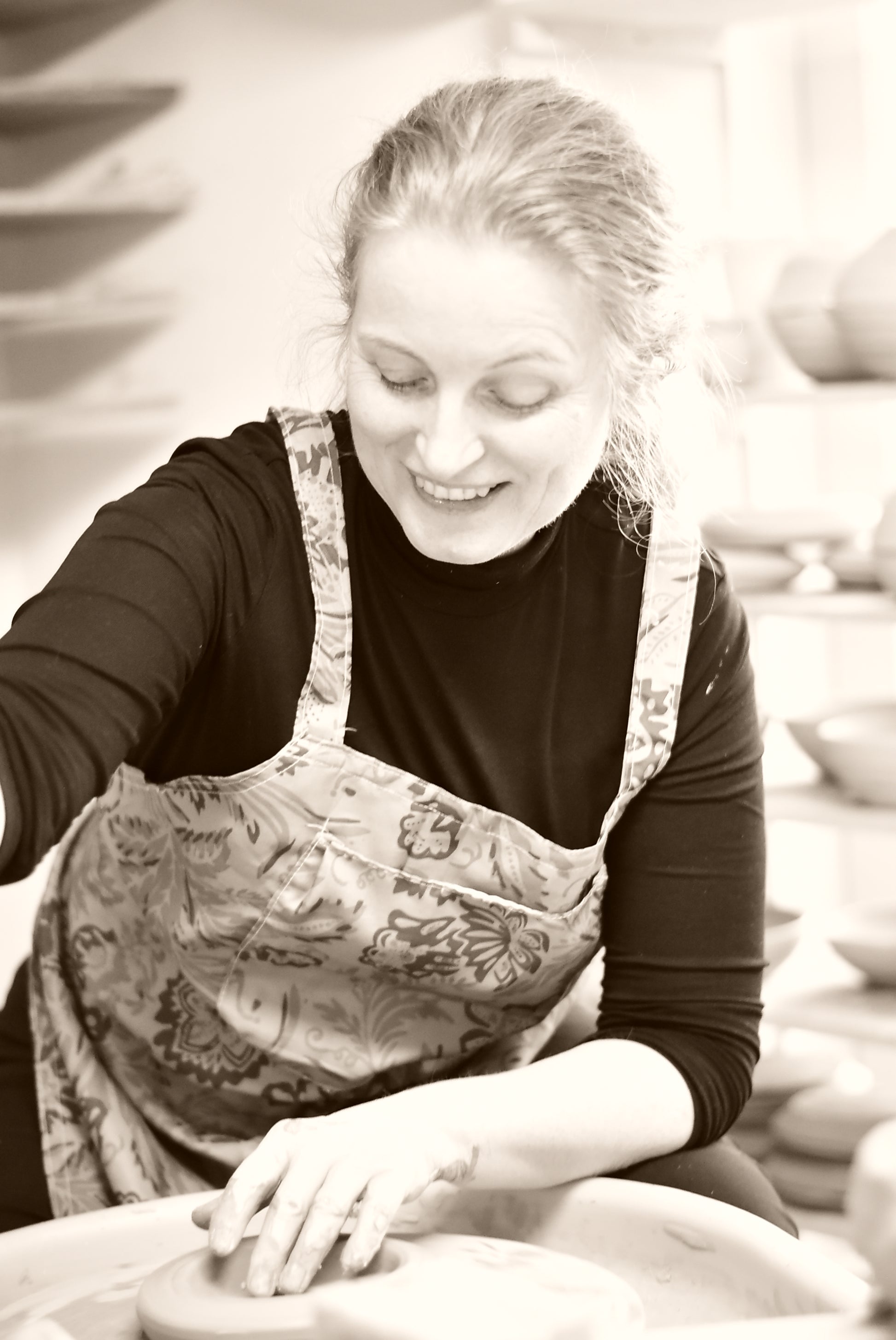 A woman smiling while working at a pottery wheel, wearing a dark long-sleeve shirt and a patterned apron in a pottery studio.