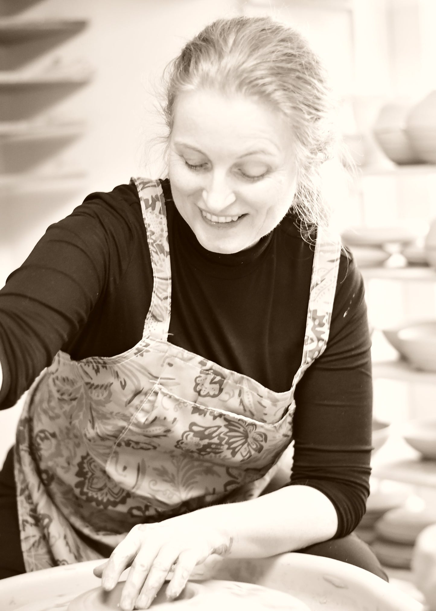 A woman smiling while working at a pottery wheel, wearing a dark long-sleeve shirt and a patterned apron in a pottery studio.