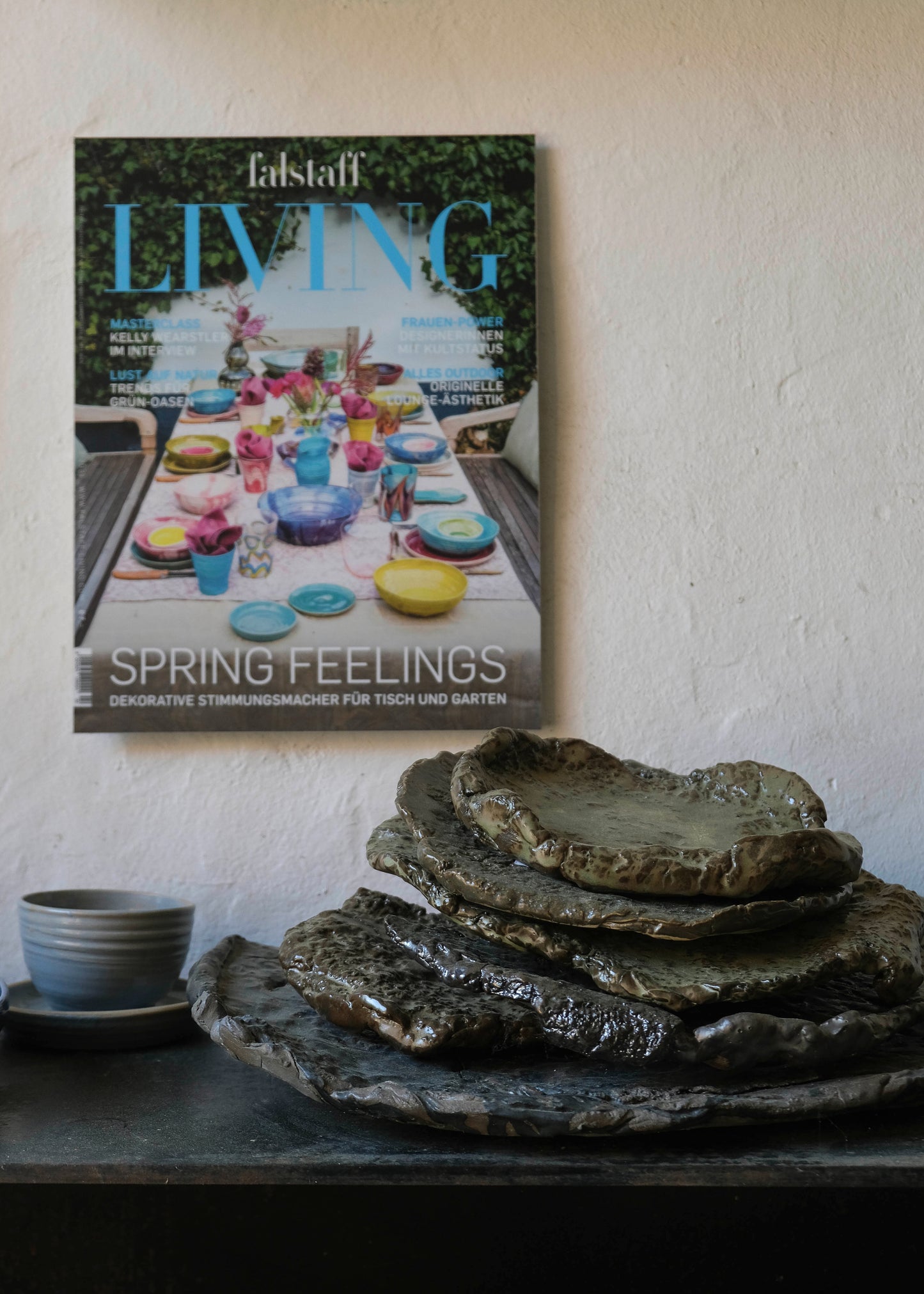 A magazine titled 'Elsstaff LIVING' with the cover story 'SPRING FEELINGS' and a colorful table setting photo, displayed on a textured off-white wall above a dark table holding stacked rustic stone plates and a small gray cup.